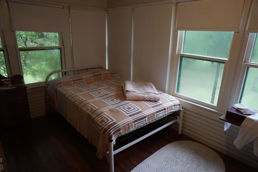 A corner room walled with windows on two sides, looking out into green trees, with a single iron bed in the middle — a sleeping porch.