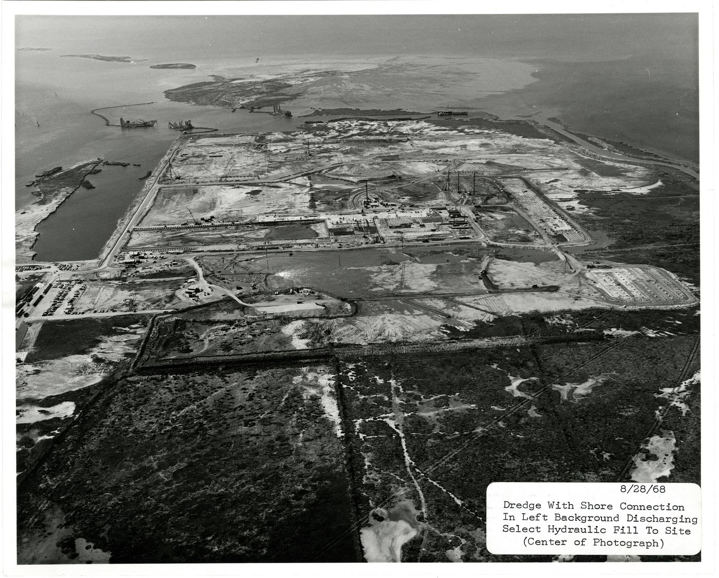 An aerial of a hydraulic dredge operation: a dredge offshore pumps bay-bottom slurry through a pipe onto a raw, staked-out shoreline, building new land above the water.