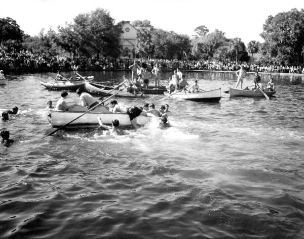 Teenage boys thrashing in the water of a bayou beside several rowboats, diving for a cross, while a large crowd lines the far shore in front of a church.